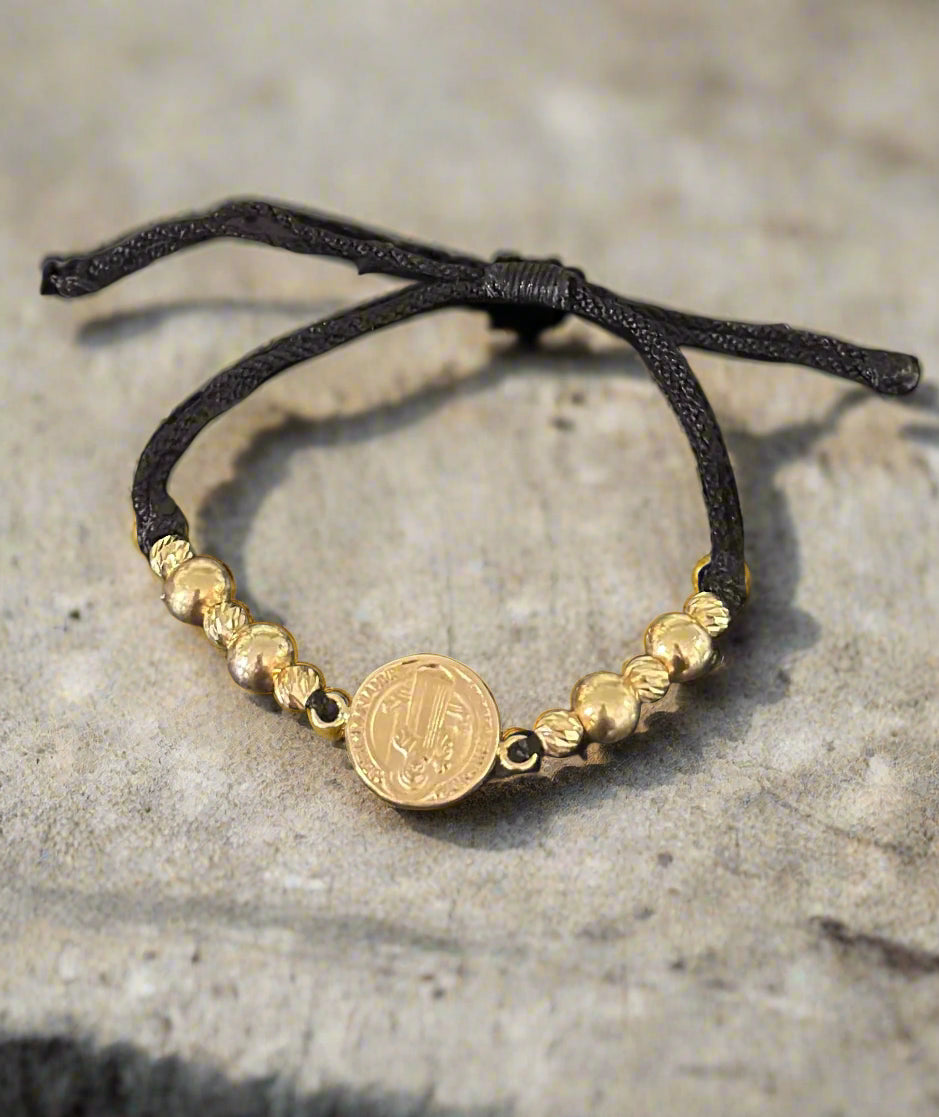Gold beaded bracelet with a a center piece of the Saint Benedict charm with diamond cut beads on a black cord against a colorful street background